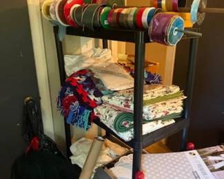 Black metal mobile ribbon storage rack with multiple spool rods
Condition: Good
Tall black metal craft organizer on wheels with many horizontal rods holding numerous ribbon spools. The rack appears used as storage in a sewing/craft space and is shown with ribbons and folded fabric on/around it.