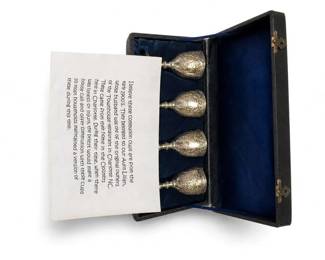 Silver-plated engraved communion cups in presentation case
Condition: Good
Four ornate silver-tone cups with engraved floral/scroll decoration in a hinged presentation case lined in blue velvet-like fabric. The object appears to be a communion or church liturgical travel/presentation set. The paper note in the box references communion cups and claims an 1800s origin, but that remains unverified from the photo alone.