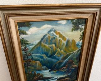 This framed oil painting depicts a dramatic mountain landscape with a snow-capped peak, lush green slopes, and a winding river in the foreground. The artwork is set in a gold-toned frame and mounted on a white wall, showcasing its vivid colors and detailed brushwork.