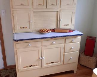 Hoosier cabinet featuring a roll-top center, a blue-and-white speckled porcelain enamel work surface, and decorative heart-and-flower stencils along the bottom doors.