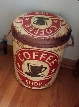 Cylindrical barrel shape featuring a cream and red distressed "Coffee Shop" graphic. Features: Equipped with a padded, removable top lid and side rope handles. Function: Doubles as a small seat/stool and an interior storage compartment.