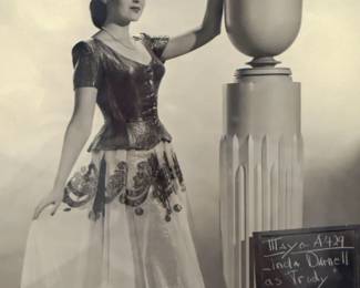 Black and white costume test photograph of a full skirted gown beside a tall decorative vase and production slate