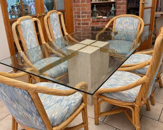 Glass-top dining table with a pale stone-look pedestal, brass-tone supports and six rattan-style armchairs upholstered in blue-and-cream patterned fabric. The sculptural base and curved chair frames suit an Art Deco Revival interior. Decorative objects in the background are separate estate items; ask at the sale about pricing and availability.