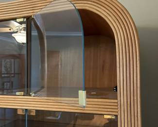 Detail of the curved rattan cabinet showing its open glass door upper shelves and brass tone hardware