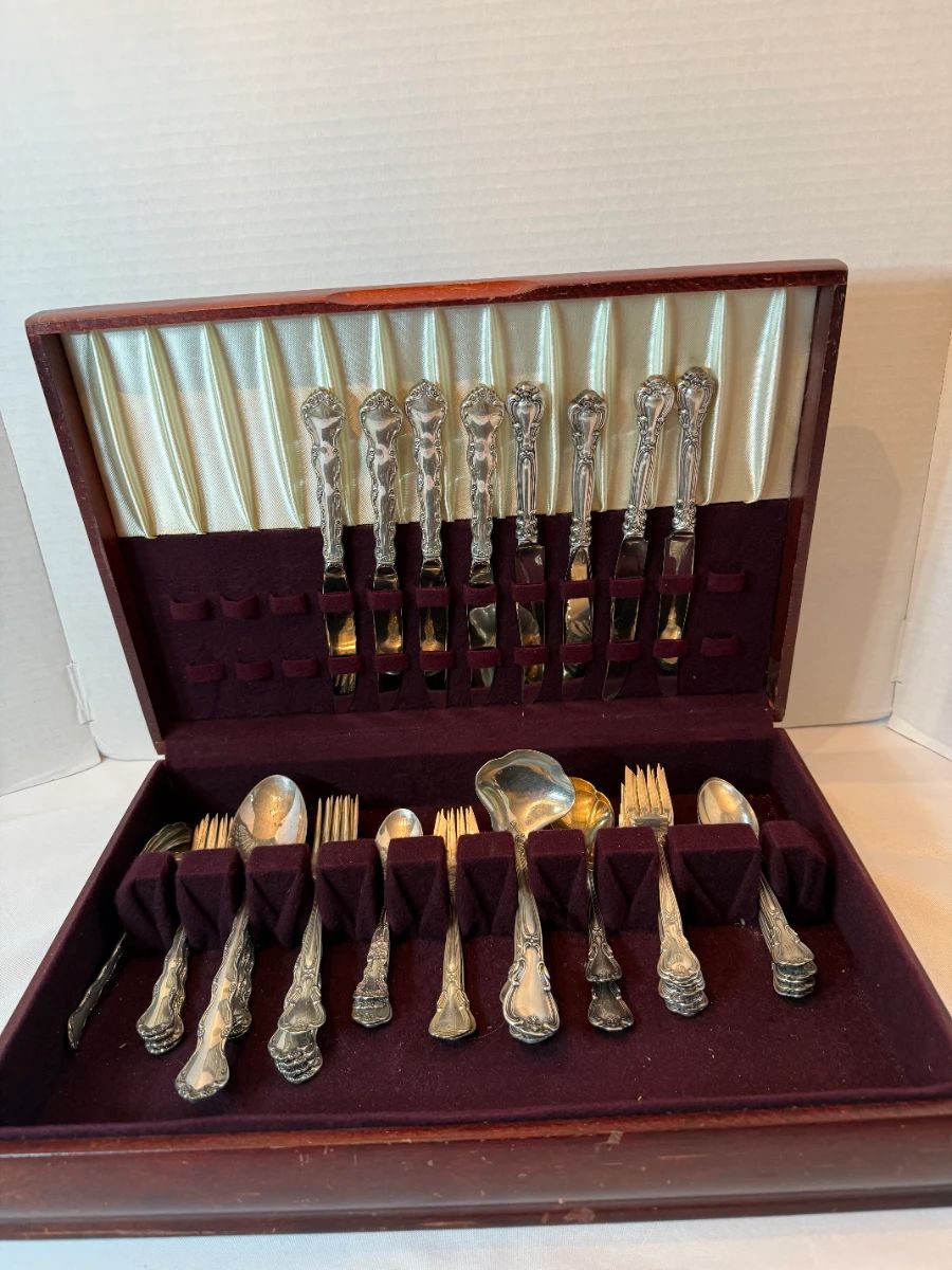 Sterling Silver Flatware Set in Wooden Chest  A sterling silver flatware set housed in a fitted wooden storage chest with a tarnish-resistant burgundy lining. The chest holds knives in the lid rack with forks, spoons, and serving pieces slotted in the base tray. The handles carry an ornate scrolled floral pattern and the pieces are bright and evenly polished. Presented ready to display or gift in its original chest.