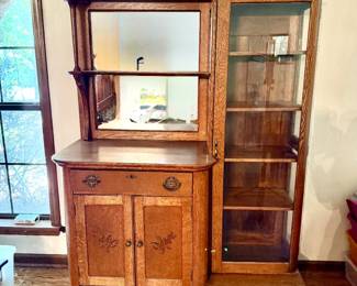 Oak Washstand with Mirror and Glass Display Cabinet A washstand with a hinged mirror in a shaped top gallery with a small side shelf, over a single drawer with brass pulls and a two-door cabinet below with carved detail on the door panels. Beside it stands a glass-front display cabinet with four wood shelves. Both pieces show a warm finish with quartersawn grain visible. Wear is consistent with age; the mirror is clear.