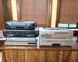 Lot of VCRs and DVD Players - A six-piece lot of home video equipment including a Sharp 4-head VHS VCR, a Symphonic VCR/DVD combo, an Emerson 4-head Hi-Fi VHS/DVD player, an Apex Digital ADV-3800 VCR/DVD combo, an Apex AD-1225 DVD player, and a Pioneer DV-260 DVD player. Several units have attached power cords visible. None of these units were tested — working condition not confirmed, sold as found. VHS playback equipment has become genuinely hard to source, which keeps demand steady from collectors digitizing home video libraries.