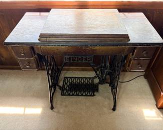 Antique Singer Treadle Sewing Machine in Oak Cabinet - An antique Singer treadle sewing machine housed in a four-drawer oak cabinet on the classic Singer cast iron base, with the "SINGER" name plainly cast into the stretcher. The wooden cabinet top and drawer fronts show significant finish loss, chipping, and honest age wear throughout. The machine head is enclosed and was not opened or photographed, so the model and condition of the machine itself are unconfirmed — working condition not confirmed, sold as found. Treadle bases alone have strong demand for repurposing into console tables, which gives this lot two audiences.