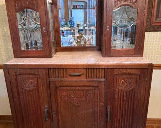 Antique Marble Top Sideboard with Beveled Mirrors An antique sideboard with a rose-toned marble top, a central beveled mirror flanked by two beveled mirrored doors set into carved frames, and a scalloped crest across the top. Below are a long central drawer and three cabinet doors, each with deep carved floral and fluted detail. The wood carries a warm reddish finish with heavy carved ornamentation throughout. A commanding period piece with a great deal of hand detail.