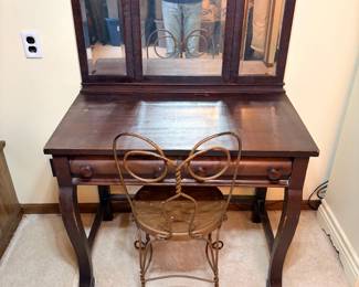 Antique Slant Desk with Trifold Mirror and Wire Parlor Chair An antique desk in a dark finish with a single drawer, shaped legs, and a hinged trifold beveled mirror mounted above the work surface. Paired with it is an antique twisted wire ice cream parlor chair with a round wooden seat and heart-shaped back, finished in a gold tone. The desk shows finish wear and scuffing across the top and edges; the wire chair shows oxidation and paint loss consistent with age. A charming vanity or writing desk lot with an unusual mirrored top.