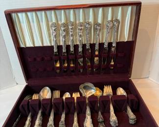 Sterling Silver Flatware Set in Wooden Chest  A sterling silver flatware set housed in a fitted wooden storage chest with a tarnish-resistant burgundy lining. The chest holds knives in the lid rack with forks, spoons, and serving pieces slotted in the base tray. The handles carry an ornate scrolled floral pattern and the pieces are bright and evenly polished. Presented ready to display or gift in its original chest.
