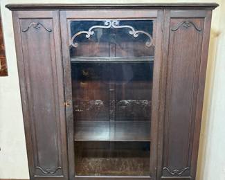 Antique Bookcase with Glass Door and Carved Panels A dark-finished bookcase with a single glass door flanked by carved side panels, topped with a pierced scrollwork gallery across the upper frame. The interior holds fixed shelving. Shaped carved detail accents the corners of both side panels and the door surround. A substantial storage piece with strong architectural presence.