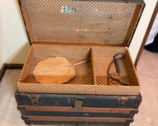 Antique Steamer Trunk with Fan An antique steamer trunk with wood slat banding, metal corner caps, and leather side handles, opening to a checkered paper-lined interior with the original lift-out tray in place. A woven hand fan with a turned wooden handle and a small metal piece are included inside. The exterior shows age wear to the finish and hardware; the interior paper is largely intact. Trunks like this get used as coffee tables and blanket storage as often as they get collected.