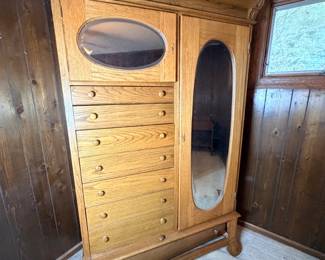 Antique Oak Chifforobe with Beveled Oval Mirrors - A tall antique oak chifforobe combining a seven-drawer bank with a full-length mirrored wardrobe door and a smaller beveled oval mirror on the upper cabinet. Carved leaf detailing accents the top corners, and the piece stands on shaped feet with a lower stretcher rail. The oak grain is prominent and the finish is warm and consistent; the mirrors appear clear with no visible cracks. A substantial antique oak wardrobe for a bedroom, and a natural companion to the marble top dresser in this sale.