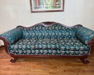 Victorian Style Sofa with Teal Floral Upholstery A sofa with a carved crest, scrolled arms, and shaped wooden feet, upholstered in a teal floral fabric with a single long seat cushion. The carved wood frame is exposed along the back, arms, and base. The upholstery is clean in the photo and the frame shows light finish wear. A statement piece for a formal front room.