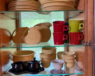Dinnerware Collection with Colored Mugs A dinnerware collection in a warm tan glaze, including dinner plates, a disc pitcher, handled bowls, nesting mixing bowls, and cups, displayed with a set of mugs in red, yellow, and green. A black covered sugar and creamer on a matching tray completes the group. The glazes are glossy and evenly colored throughout. A bright, functional set with good shelf appeal.