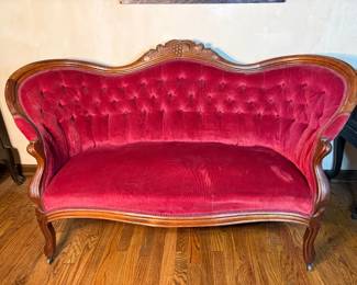 Victorian Tufted Settee in Red Velvet A settee with a serpentine carved crest, deep button tufting across the back, and scrolled arms flowing into cabriole legs. The upholstery is a rich red velvet, full and even through the tufting, set against a warm wood frame. The carved crest features a floral and foliate motif at the center. A striking centerpiece for a parlor or entry.