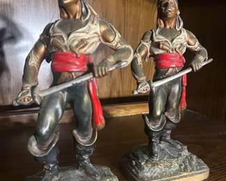 Pair of Paul Herzel Antique Pirate Figurines with Red Sashes Two cast figurines depicting standing warriors in flowing trousers with red sashes at the waist, each holding a staff or pole arm, mounted on rectangular bases. The finish shows a dark patina with painted red and flesh-tone highlights, and both show honest age wear with some paint loss. The metal content is not confirmed from the photo and the pieces were not checked for foundry marks — verify before describing the material. A striking decorative pair with good shelf presence.