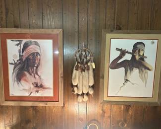 Pair of Framed Native American Portrait Prints with Dreamcatcher Two framed prints depicting Native American subjects — one a headband-wearing portrait, the other a figure playing a flute — in coordinating wood frames with mats. A feathered dreamcatcher with hanging plumes is included between them. Both prints appear clean behind glass; artist signatures and edition markings were not legible in the photo and should be checked at the lower margin. A coordinated three-piece wall grouping.