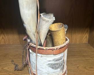 Birch Bark Bucket with Hunting Horn Calls - A birch bark bucket wrapped with a leather-laced band, holding a grouping of hunting horn calls along with a feathered wing piece. The horns range in size and tone from pale to deep amber, and the bark surface shows its natural white and gray markings. The leather lacing and carry cord are intact. A distinctive display grouping for a hunting room, den, or cabin.