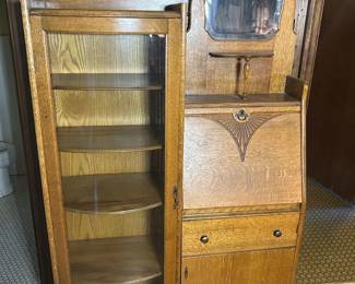 Antique Oak Side-by-Side Secretary Bookcase An antique oak secretary bookcase in the side-by-side configuration, with a curved glass door enclosing four shelves on the left and a drop-front writing desk over a single drawer and lower cabinet on the right. The drop front carries an incised fan-and-scroll carved motif, and a small beveled mirror is set into the upper gallery. The oak grain is prominent and the finish is warm; hinge, lock, and glass condition should be confirmed in person. A desirable form for antique oak buyers and one of the stronger furniture pieces in this sale.