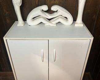 White Cabinet with Abstract Sculptures A two-door white cabinet with brushed pulls, shown with a white abstract sculpture depicting two interlocking figures and a pair of tall white figures on top. The cabinet is clean and serviceable with light wear at the edges. The sculpture's material and any signature were not verified in the photo and should be checked. A practical storage cabinet with a modern decorative grouping.