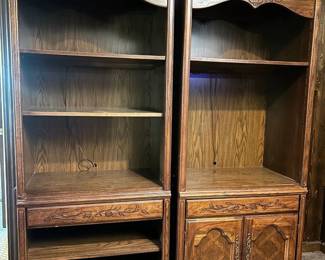 Pair of Tall Wood Bookcases with Lower Storage Two matching tall bookcases with arched crown tops and adjustable upper shelving. The left unit has a single drawer at the base; the right unit has a two-door cabinet with raised panel fronts. Both show a medium wood finish with visible grain, and there is light wear along the shelf edges and base. A matched pair like this works well flanking a doorway, fireplace, or television.