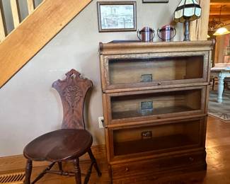 Antique carved wood sewing or spinning chair along with an antique oak 3 glass stacked book shelves and a bottom drawer. Artwork on wall features Nantuckett Island.
