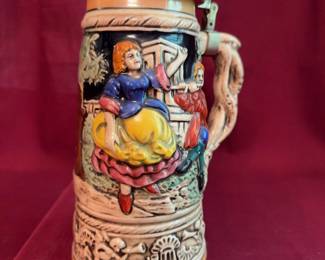 Ceramic beer stein featuring a detailed relief scene of a dancing couple and alpine chalet, fitted with a pewter lid and ornate handle. Measures approximately 9.5 inches tall. The base includes windup music box components.