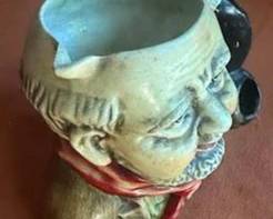 Hand-painted ceramic character mug depicting a bearded man wearing a red scarf, featuring a pipe as the handle. The base is marked 'Austria.' The mug displays detailed facial features and textured hair.