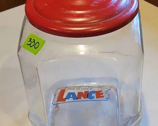 Vintage Lance glass cookie jar featuring a red metal lid and original logo label. The jar stands approximately 12 inches tall. Displays some signs of surface wear on the lid. An iconic collectible for advertising or kitchenware enthusiasts.