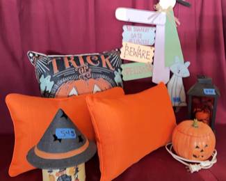 A ceramic jack-o-lantern lamp with a plug, a painted metal lantern with a Halloween scene, a fabric Trick-or-Treat pillow, two orange accent pillows, a painted Halloween-themed jar with a lid, and a wood witch decorative sign.