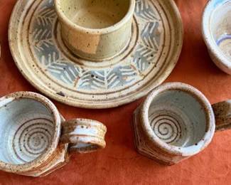 Wheel-thrown studio pottery vessels and plates, including cups, bowls, a platter, and a plate with a central dipping section. Several pieces are signed or marked, including signatures such as 'Ellie' and others with makers' marks. Glazed finishes vary throughout the collection.