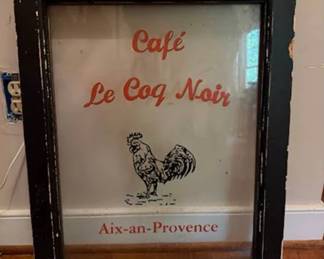Framed glass sign featuring red lettering 'CafÃ© Le Coq Noir' and 'Aix-an-Provence' with an illustrated rooster. Set in a black wooden frame with visible paint wear. Likely used as decorative or advertising signage.