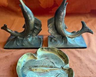 Brass sailfish-form bookends and a brass ashtray featuring a relief fish design. The bottom of the ashtray is marked with a round maker's-stamped logo and numerical code J-10. 