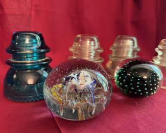 Glass insulators in different shapes and colors, and two decorative glass paperweights. The insulators vary in color, including clear and blue-green, some marked with manufacturer names. Paperweights feature embedded colored designs and bubbles.