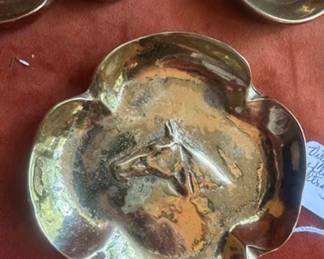 Brass ashtrays, each with four-lobed edges and central bird or horse relief designs. Bottoms are marked with 'KM' and numbered stamps. One ashtray remains in protective wrapping. Well-defined decorative metalwork with maker's mark visible.