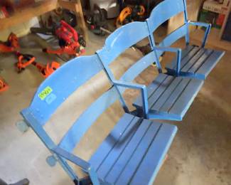 Row of three connected stadium seats with blue painted wood slats and metal frames. Seats are attached to wooden blocks for stability. Armrests are included at each end and between seats. Visible surface wear and paint loss present.