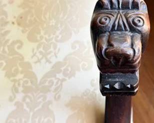 Upholstered armchair featuring wood arms with detailed carved lion head finials. Upholstery is a cream damask with visible spotting and some wear. Classic high back design with nailhead trim detailing.