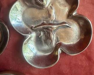 Brass ashtrays, each with four-lobed edges and central bird or horse relief designs. Bottoms are marked with 'KM' and numbered stamps. One ashtray remains in protective wrapping. Well-defined decorative metalwork with maker's mark visible.