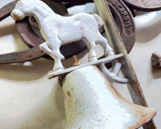 Cast iron wall-mountable bell featuring a horse figure and wheel detail. Includes several used, rusted metal horseshoes. Both bell and horseshoes show oxidation and surface wear.