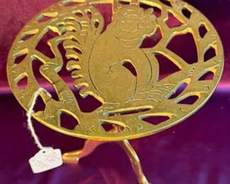 brass trivet with a cutout design featuring a squirrel sitting on a branch, supported by a three-legged stand. The trivet is decorative and functional, suitable for protecting surfaces from hot pots or dishes. 