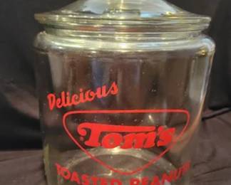 Clear glass jar with lid featuring red 'Delicious Tom's Toasted Peanuts' branding and 'An American Tradition' slogan. Jar stands approximately 10 inches tall with lid.