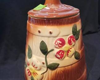 Stoneware canister featuring a brown glaze and hand-painted floral relief design, complete with a matching lid. The body and lid show some paint wear. The canister measures approximately 10 inches tall.
