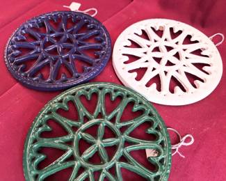 Round cast iron trivets in green, blue, and white finishes. Each features an ornate openwork starburst design and a hanging loop. 
