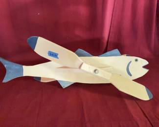 Painted wood whirligig in the shape of a fish with propeller-like fins. Features blue painted accents on the fins, tail, and face details. Designed for outdoor decorative use.