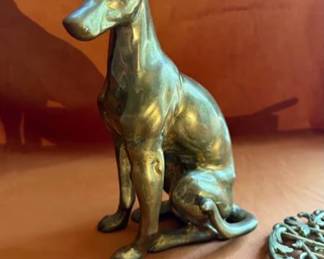 Brass dog figurine with fine sculptural detail, accompanied by brass trivets in ornate, monogram, and scroll designs. 