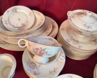 Noritake porcelain dinnerware set featuring a pink and green floral design with gold trim. Includes dinner plates, salad plates, soup bowls, cups with handles, a sugar bowl with lid, and a creamer. Items show light wear. Visible Noritake backstamp on several pieces.