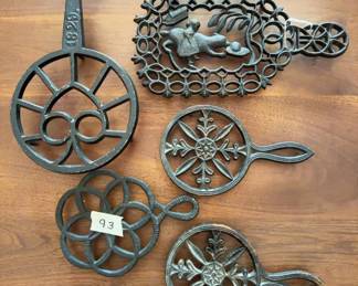 Cast iron trivets, including various designs such as a British coat of arms, figural motifs, a round floral pattern, and a rectangular tray with hunting imagery. Some pieces have visible rust and general surface wear. Several trivets are marked with foundry names or production locations on the reverse.