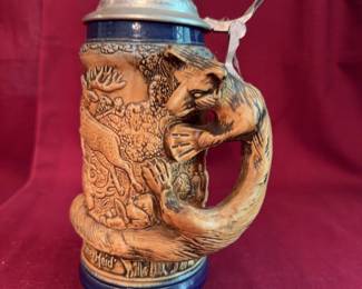 Ceramic German beer stein featuring detailed relief scenes of forest animals and hunting motifs, with phrases "Die grÃ¼ne Heid" and "Des Jaeger's Freud". Includes a pewter lid with thumb lift and a handle shaped like a fox. The bottom is marked.